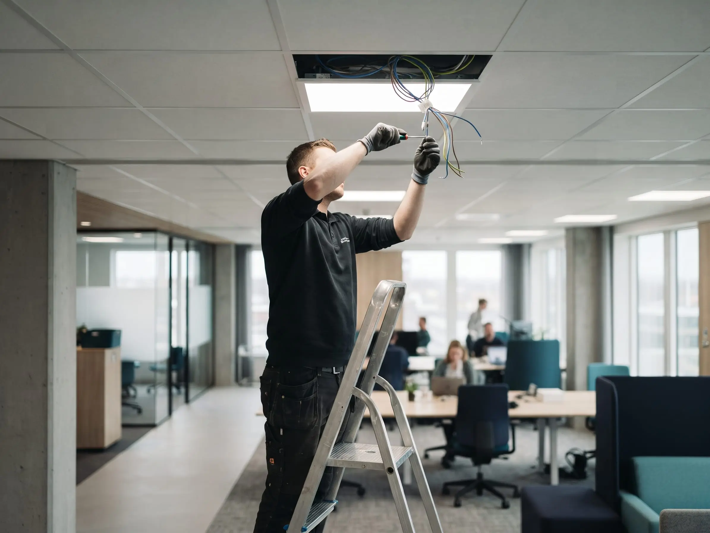 Técnico con guantes, subido en una escalera de aluminio, conecta los cables de un panel LED en el cielo raso de una oficina, con personas trabajando al fondo