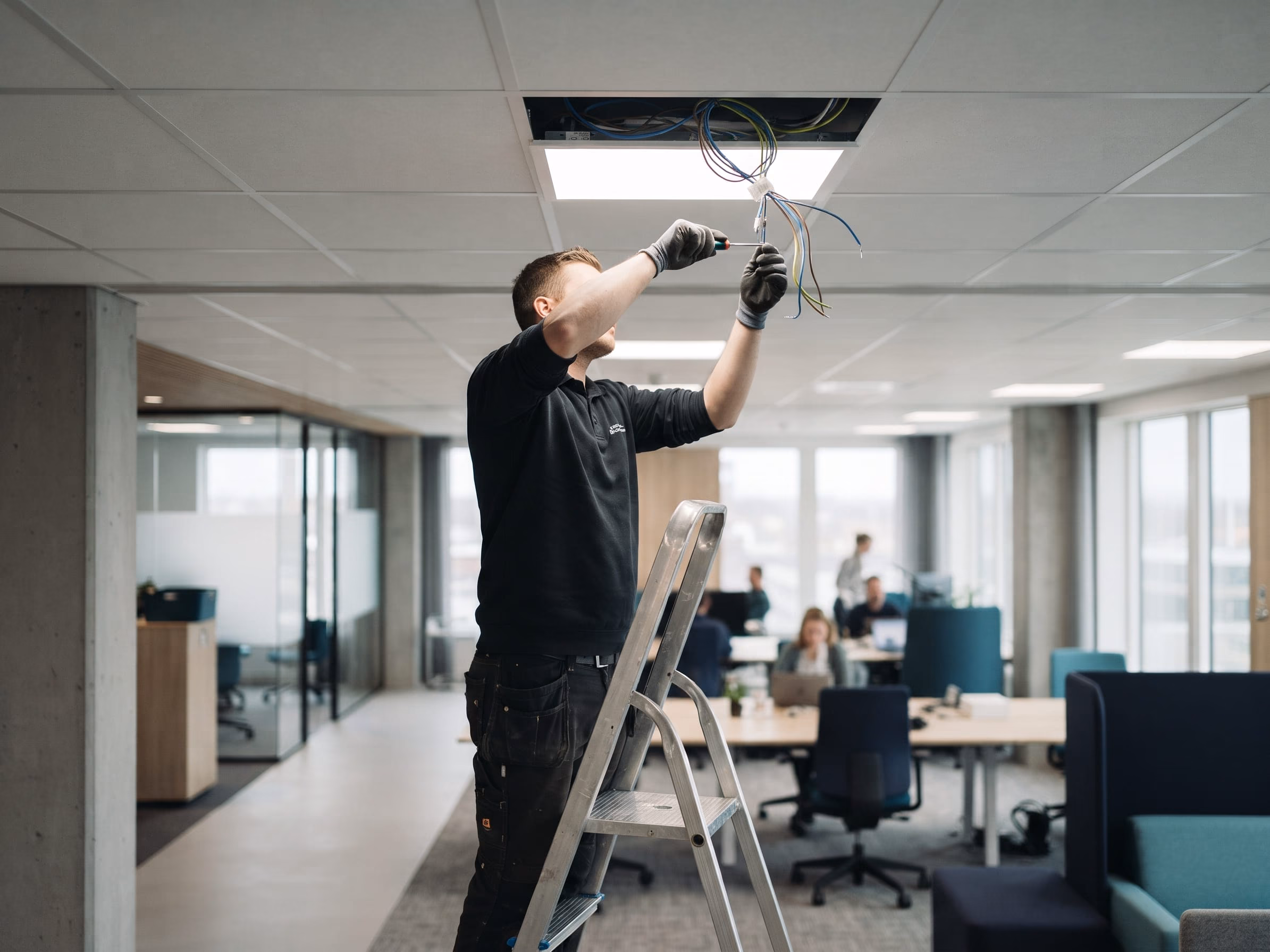 Técnico con guantes, subido en una escalera de aluminio, conecta los cables de un panel LED en el cielo raso de una oficina, con personas trabajando al fondo