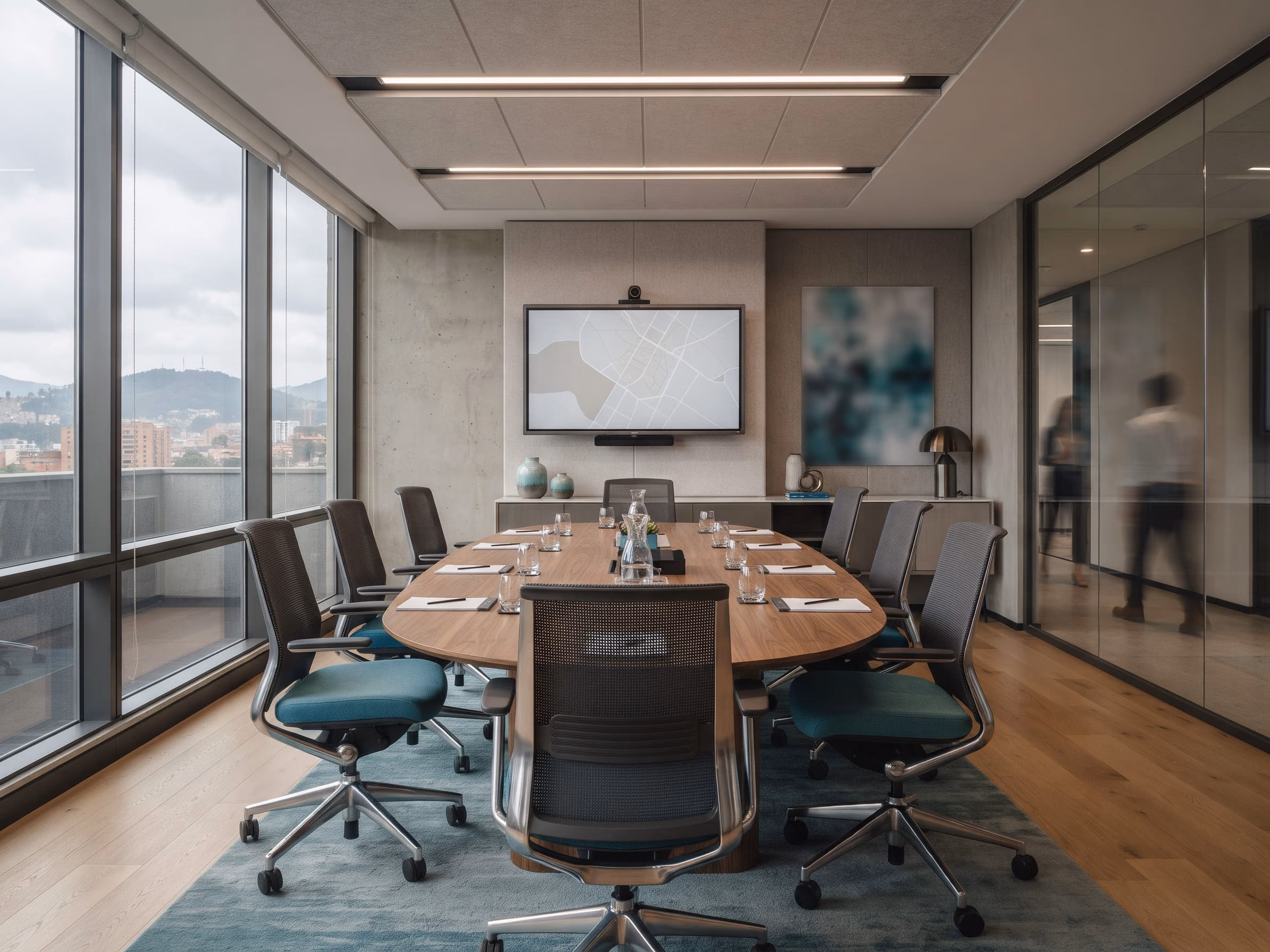 Sala de juntas con mesa ovalada de madera para diez personas, sillas de malla con asiento azul, una pantalla en la pared y ventanales hacia la ciudad