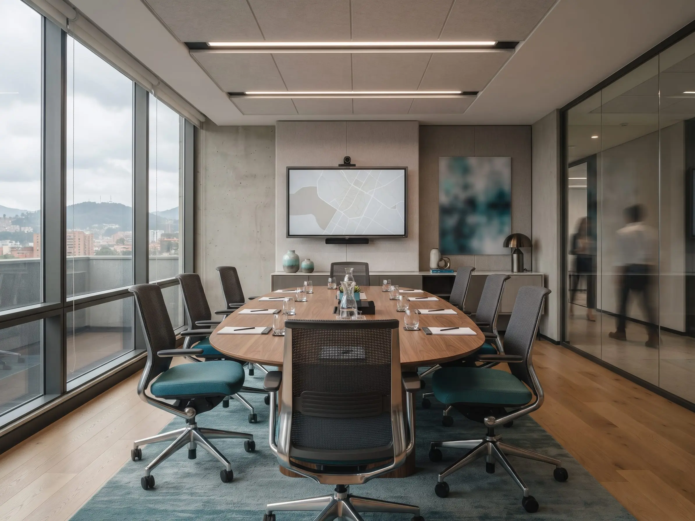 Sala de juntas con mesa ovalada de madera para diez personas, sillas de malla con asiento azul, una pantalla en la pared y ventanales hacia la ciudad
